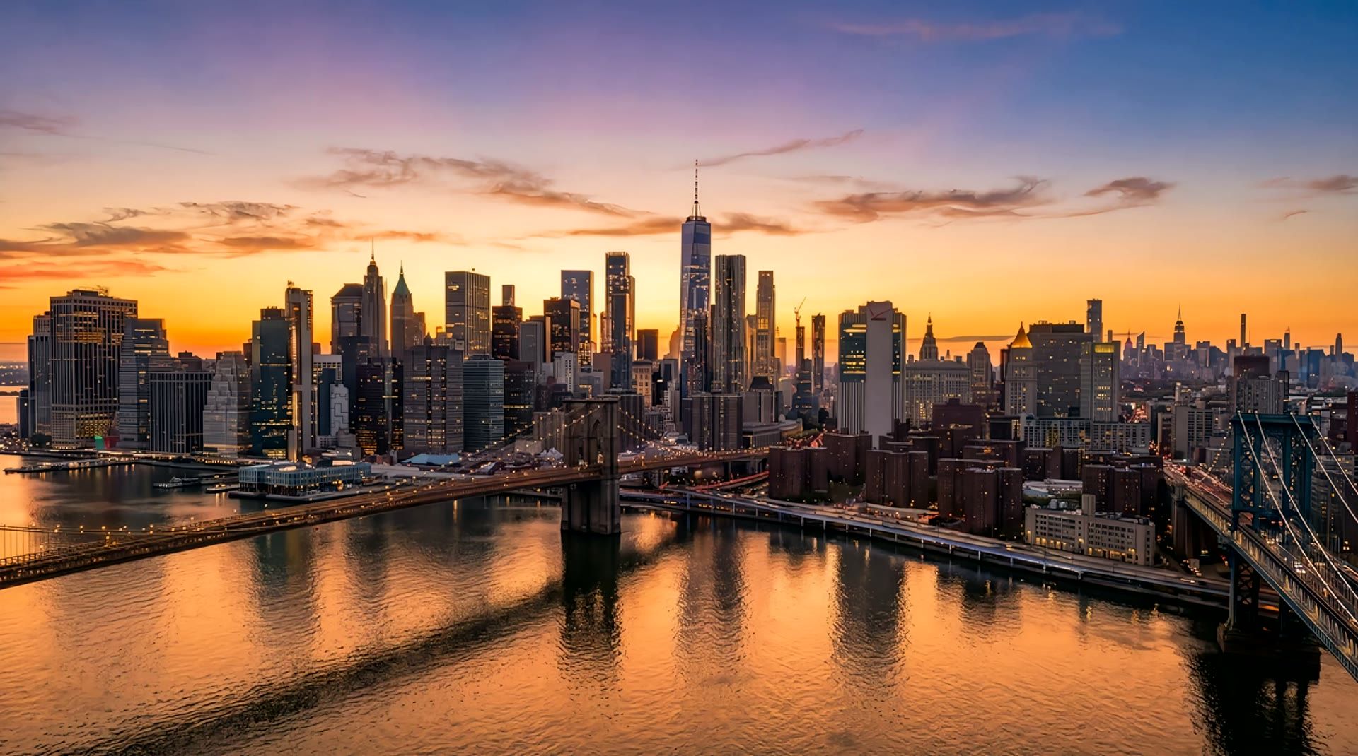 Lower Manhattan at sunset seen from above the East River, the sky turning from orange to night as the towers and bridges light up